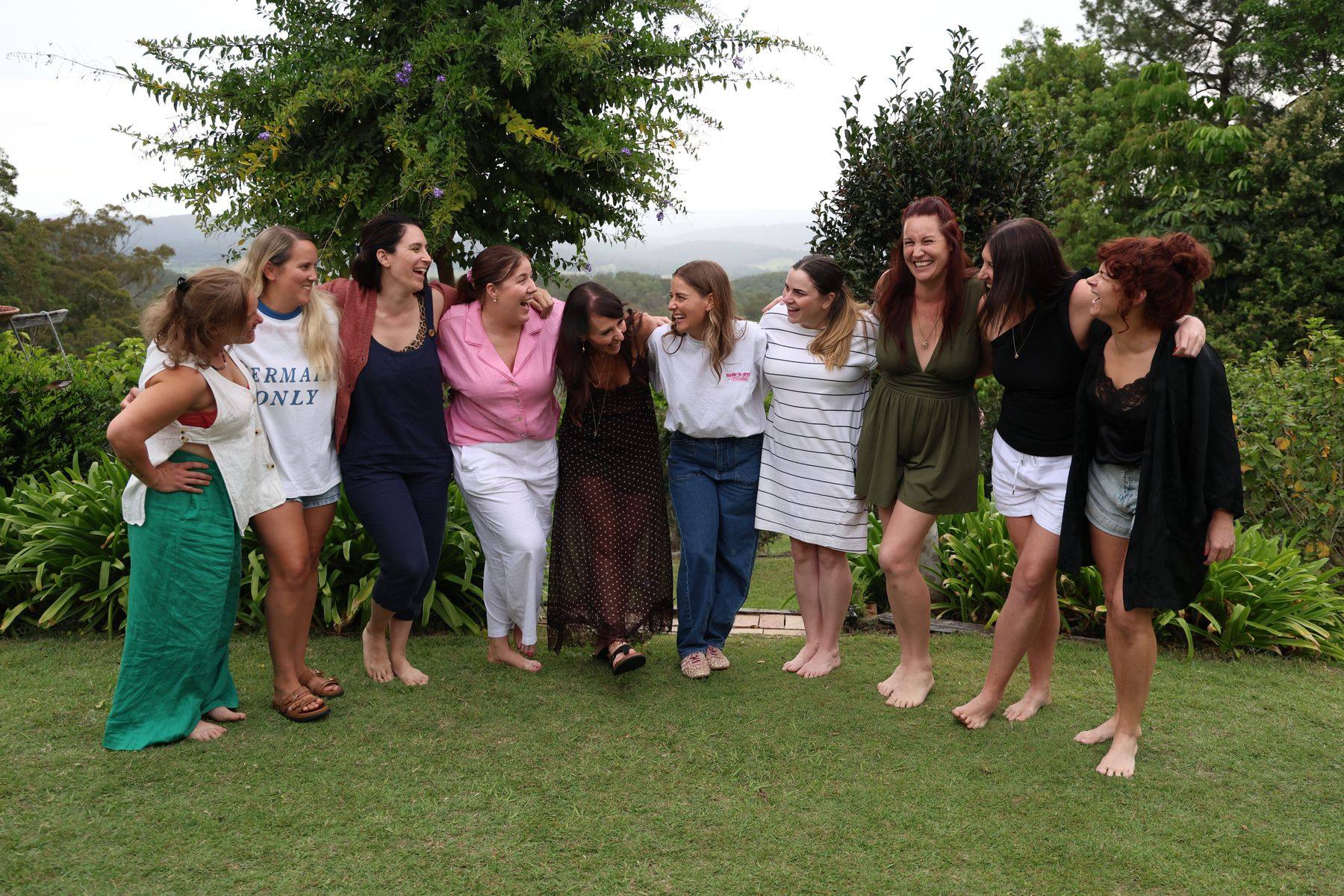 Ten women from the RECLAIM retreat standing arm in arm on the lawn, laughing, with the hinterland behind them